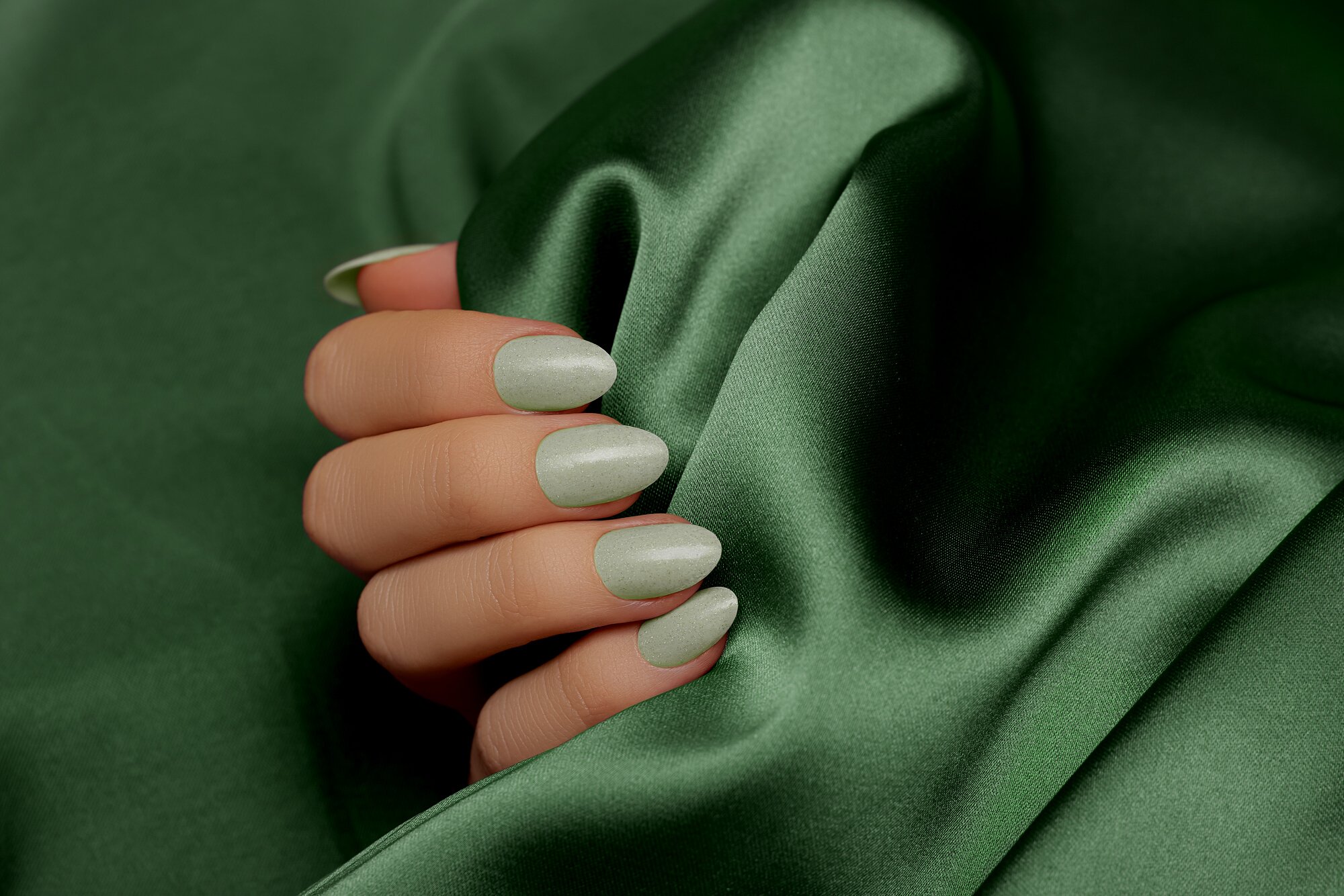 Female hand with green nail design. Light green glitter nail polish manicure on green fabric